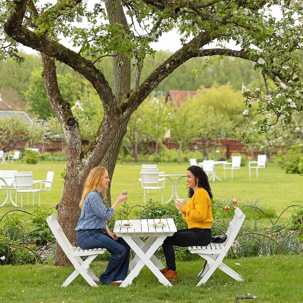 Helena och Olida sitter vid ett vitmålat bord som står under ett träd. I bakgrunden syns fler bord i trädgården.