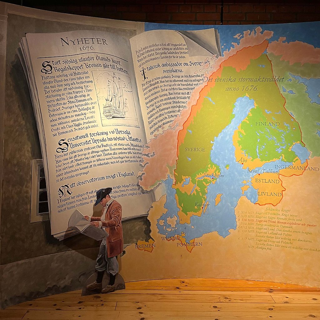 En man i historiska kläder läser en tidning. Bakom syns en jättestor bok och en karta.