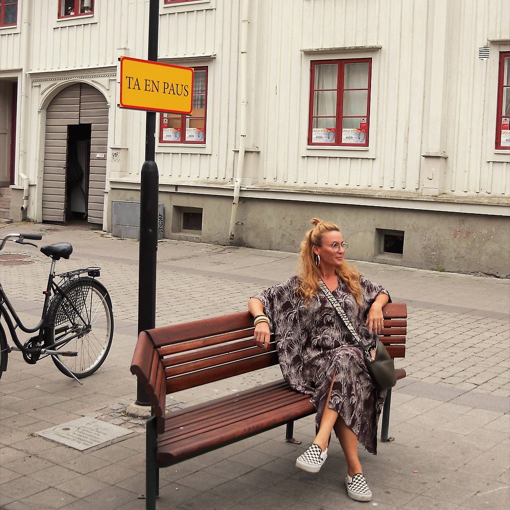 Marika Axén sitter på en parkbänk mitt i stan. Ovanför bänken finns en skylt med budskapet Ta en paus.