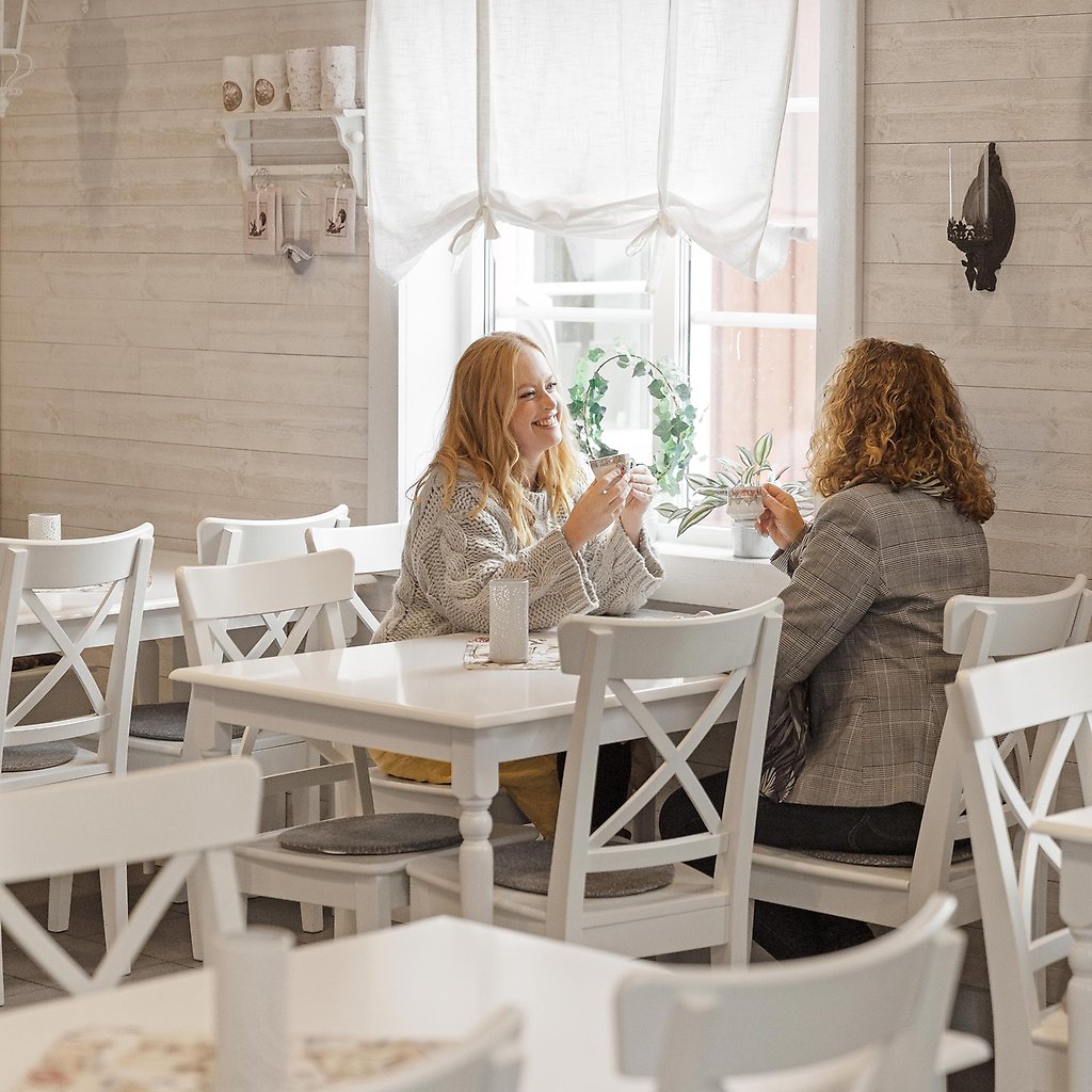 Helena och Marija sitter med kaffekoppar i handen vid ett vitt bord inne i ett kafé.