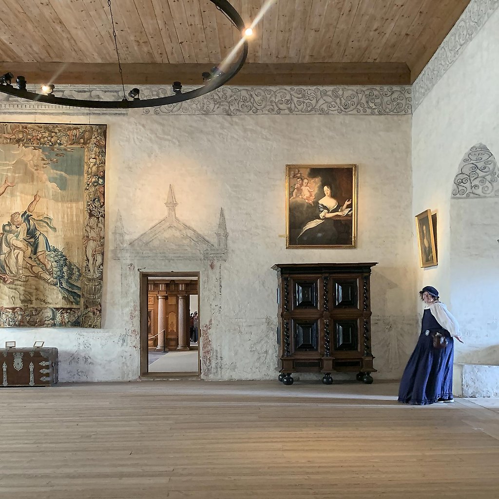En slottssal med tavlor på väggarna. Till höger står en person i historiska kläder.