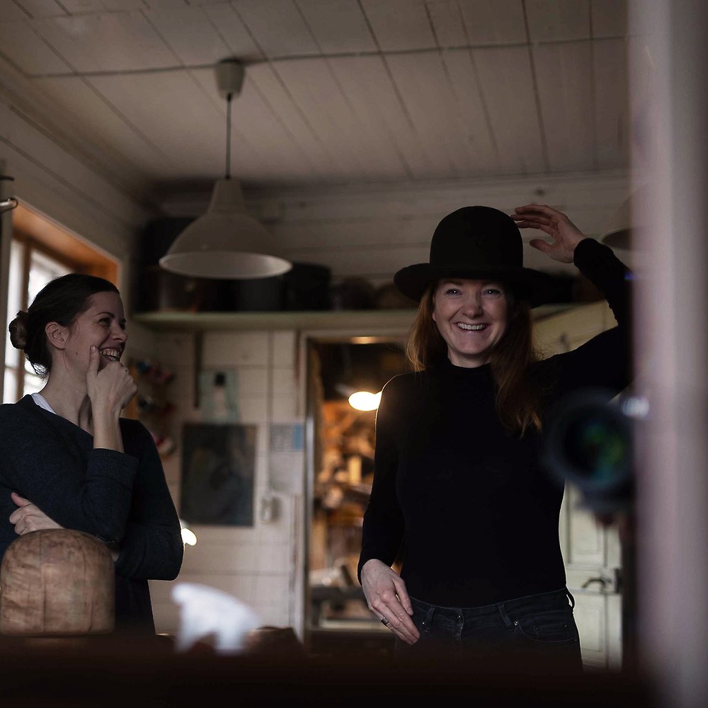 En leende Sara provar en hatt. Bredvid står en annan kvinna och ler.
