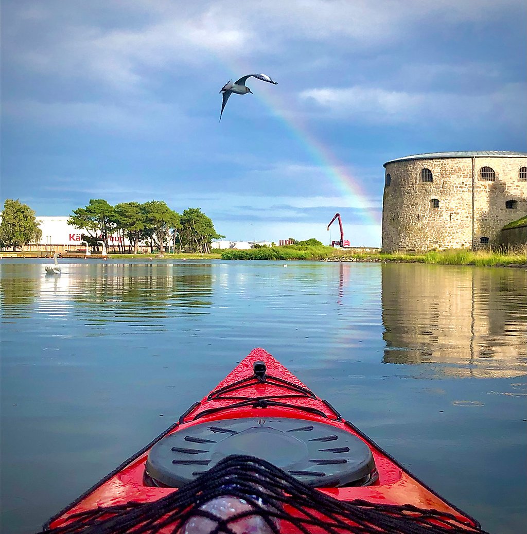The front of a kayak in front of Kalmar castle. A rainbow and a bird can be seen in the sky.