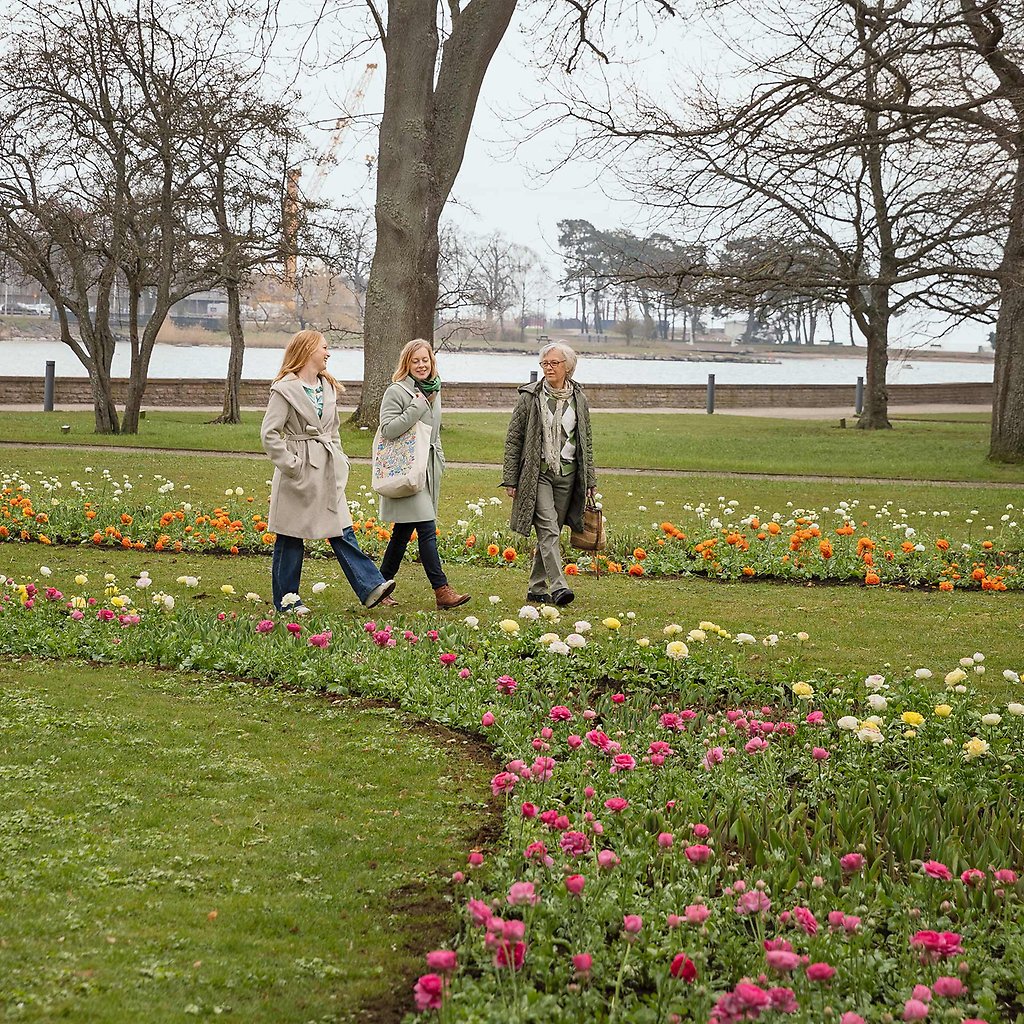 Tre personer strosar i en park, omgivna av färgglada blommor.