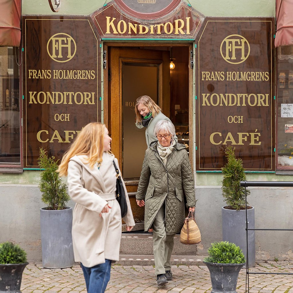 Helena, hennes mamma och syster kliver ut genom en dörr som är omgiven av bruna skyltar i äldre stil som det står Frans Holmgrens Konditori och Café på.
